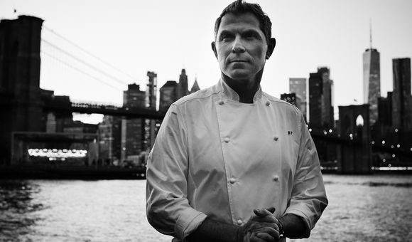 A black and white photograph of Bobby Flay in his signature white chef’s coat, with his initials, BF, embroidered on the front.