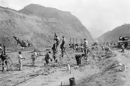 Panama Canal construction in 1913 showing workers drilling holes for dynamite in bedrock, as they cut through the mountains of the Isthmus. Steam shovels in the background move the rubble to railroad cars.