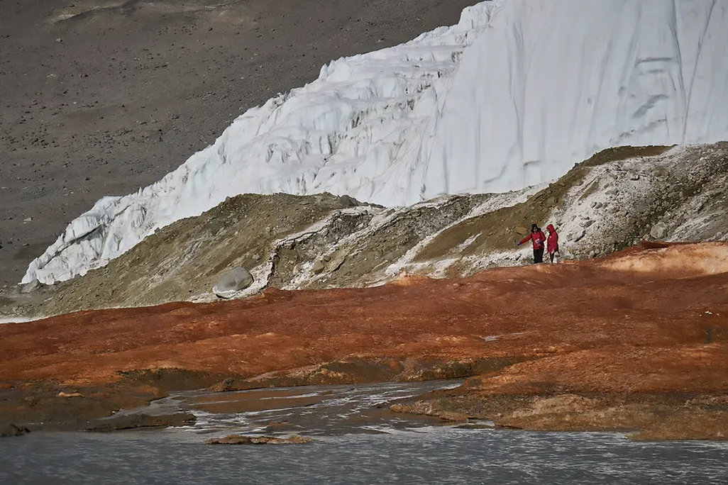 a landscape with ice, earth and red-stained ground, and two researchers