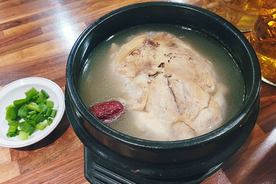 Closeup of a bowl of soup with a large piece of chicken, alongside a small dish of chopped green onion.