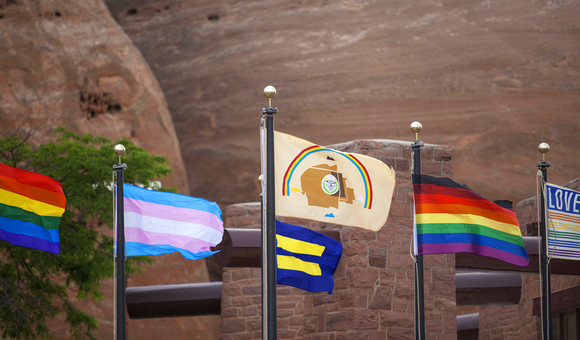 Various Pride flags (rainbow, transgender, equality, LGBTQ+ people of color, and love rainbow) displayed alongside the Navajo Nation flag in Window Rock, Arizona. (Photo by Pamela J. Peters (Diné)/Táchii’nii Photography, used with permission) 