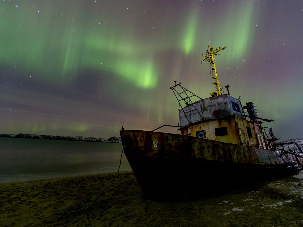 An image of a the northern lights in the sky. The photo also shows a dilapidated ship sitting on land next to water.