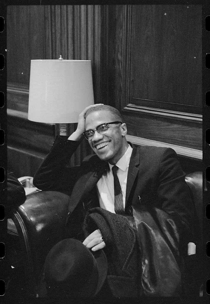 Malcolm X, a tall thin man with glasses, wears a suit and tie and puts leans his head against his arm as he laughs