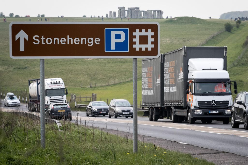 A busy road is in the foreground, with trucks and cars, and a large sign pointing to Stonehenge; the iconic stone structures are visible in the background