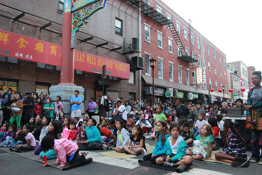 A crowd on a city street includes children seated on mats in the foreground and adults standing, facing a stage out of frame. Red lanterns hang across the street in the background.