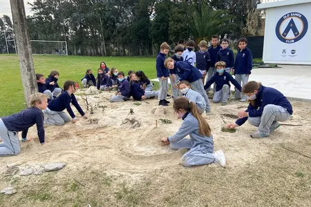 Students cluster in groups and individually around a sandy area, adding and refining the community map they are building out of sand, leaves, and sticks.