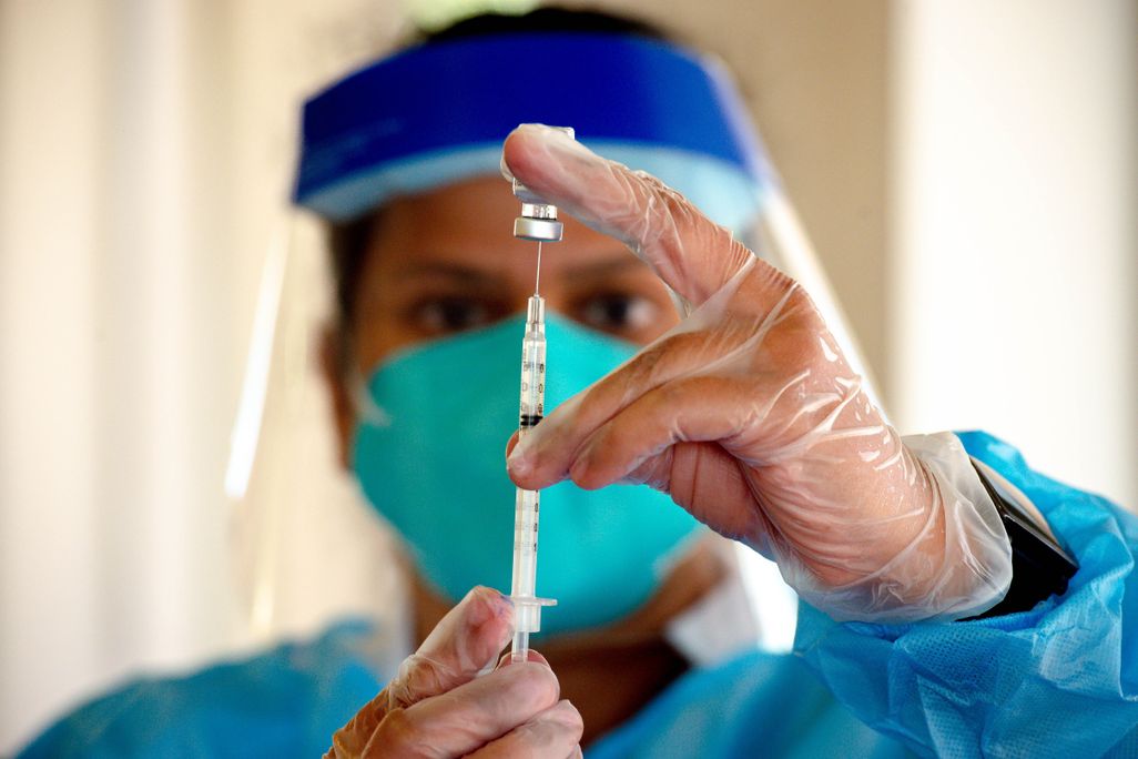 A pharmacist holds a vial of vaccine and a syringe in front of their face