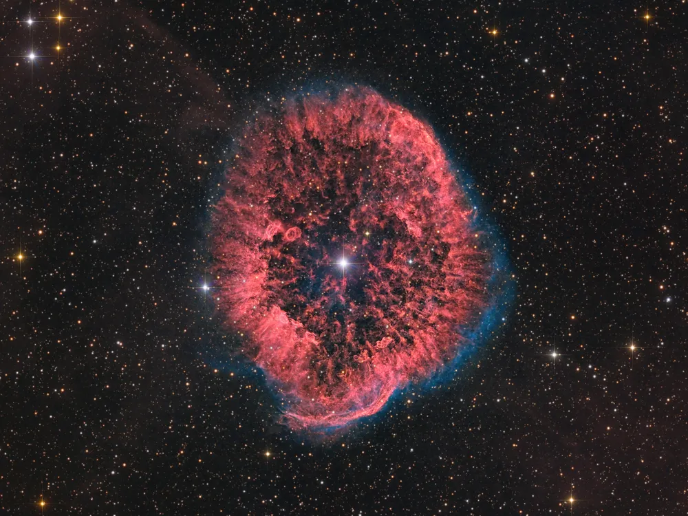 a red billowing ring of cosmic gas and dust surrounds a bright star with other stars appearing as several pinpricks of light in the background