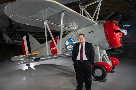 A white man in his 30s stands in front of a restored biplane displayed on a polished concrete floor inside a museum. He is wearing glasses and a dark suit with a white shirt and burgundy tie.