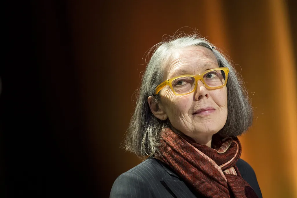 A close-up shot of an older woman with a bob hair cut wearing yellow glasses and a maroon scarf