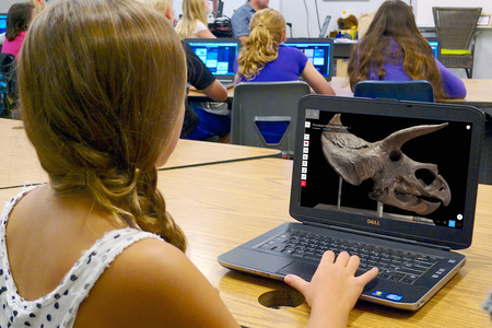 Girl in a classroom using a laptop computer that shows a dinosaur skull on its screen. Several other students work at desks with laptops in the background.