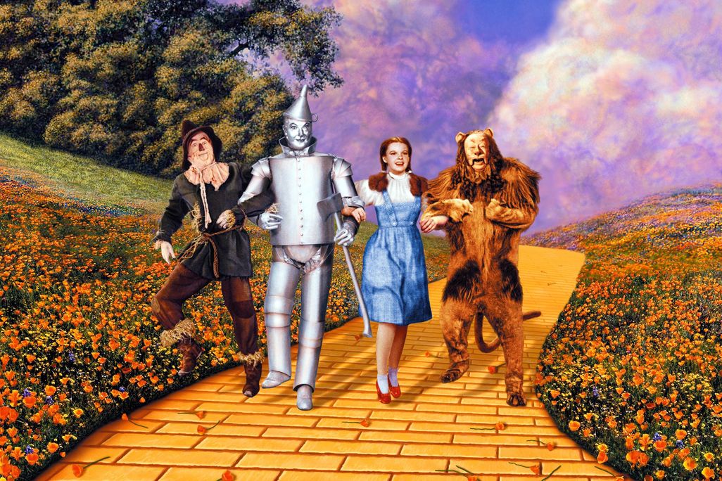 Four people, a scarecrow, Tin Man, Dorothy and a lion, walk arm in arm down a yellow brick road 