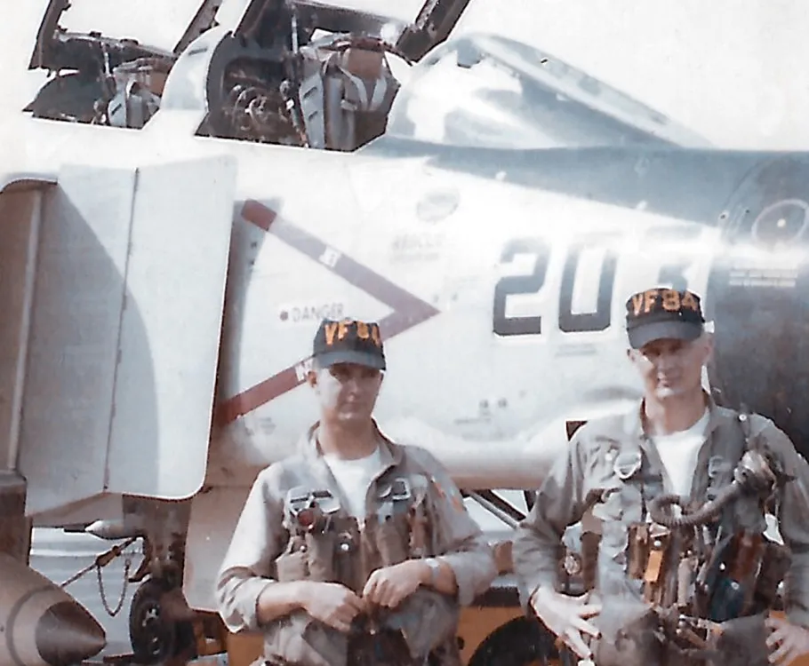 Two U.S. Navy men in uniform--a pilot and an aviator — stand in front of the cockpit of an F-4B Phantom II military jet aircraft.