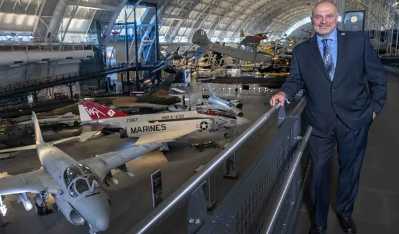 A white man in his mid-50s stands on a mezzanine overlooking several military jet airplanes parked on the concrete floor of a large hangar. The man is smiling and wearing a dark gray suit, blue shirt, and tie.