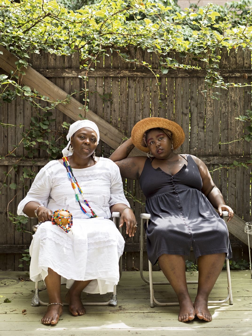 A portrait of two Black women seated outside, side-by-side, facing the viewer; one wears a white dress and the other wears a black dress and a sunhat