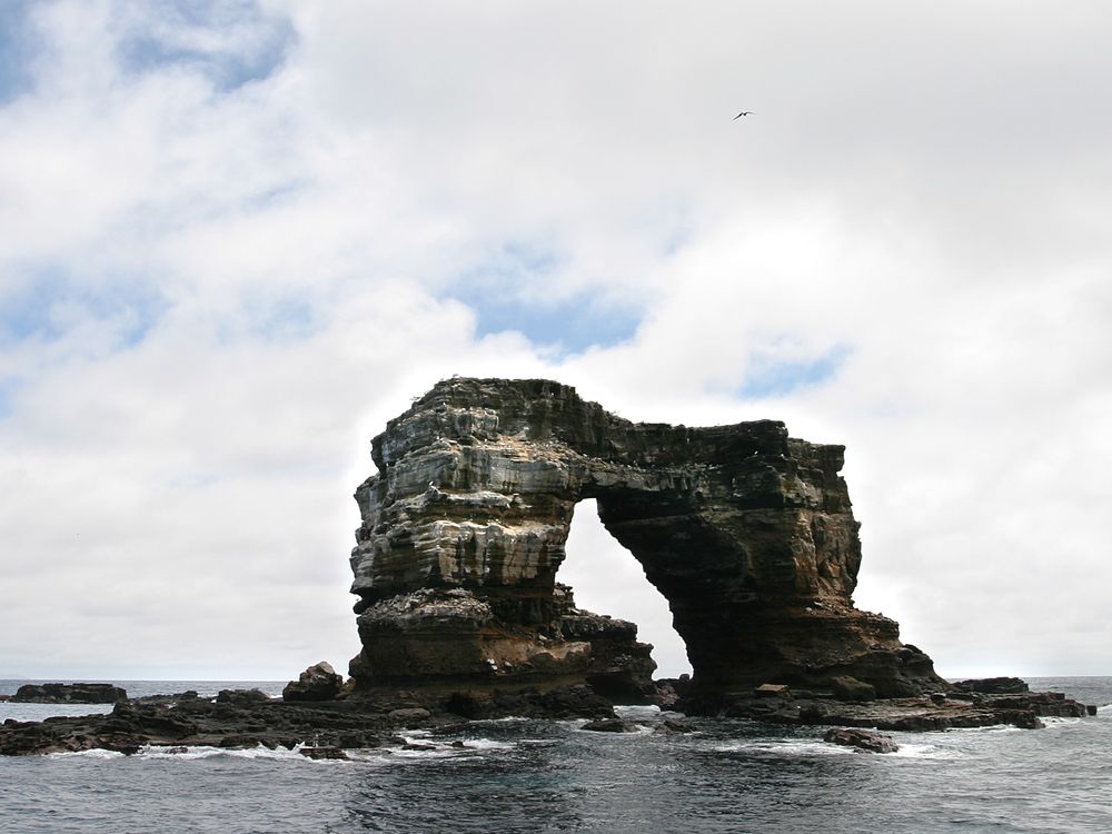 A photograph of Darwin's Arch in the Galàpagos Islands. The rock formation is two pillars attatched by a natural rock bridge. It stands itself in the ocean with a cloudy sky in the background.