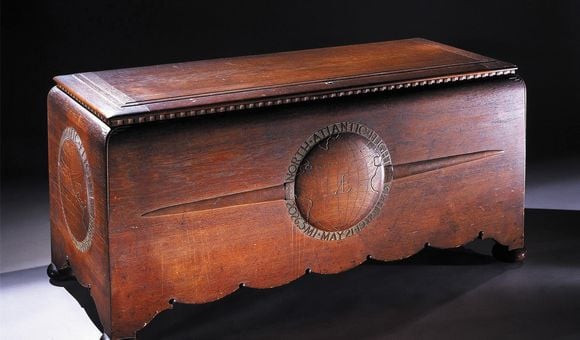 A large hand-carved teakwood chest with scalloped edges and four round feet. The chest appears to be stained a rich reddish brown. The front panel on the chest has a bas-relief globe pattern flanked by a pair of airplane wings.