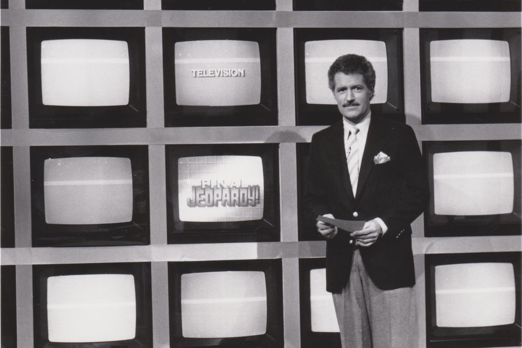 A black and white image of a young Trebek, a white man in a suit and tie with dark hair and a mustache, standing in front of a grid of TV screens