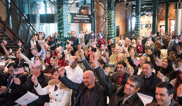 A group of people sit in chairs at the History Center raising mini American flags during a Citizenship ceremony