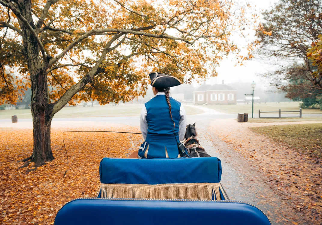 A horse-drawn carriage travels along a leaf-covered road in Colonial Williamsburg during autumn. A driver in period clothing sits at the front beneath golden fall foliage, with historic buildings visible through the morning mist.