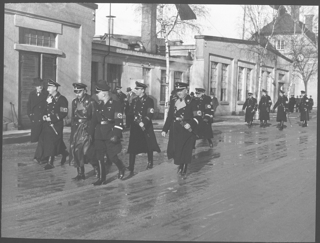 The commandant’s office building (in the top right corner) and the still-standing workshop building (in the middle and on the left) appear in this photograph of Nazi leaders, including Heinrich Himmler, visiting Dachau in January 1936.