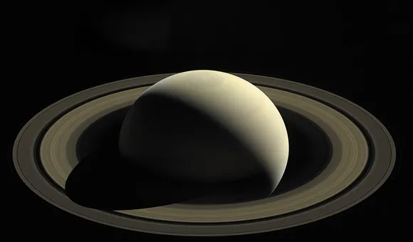 The smooth surface of a round planet is partially illuminated by the sun; the planet's other half is in shadow. The planet is surrounded by thousands of rings.