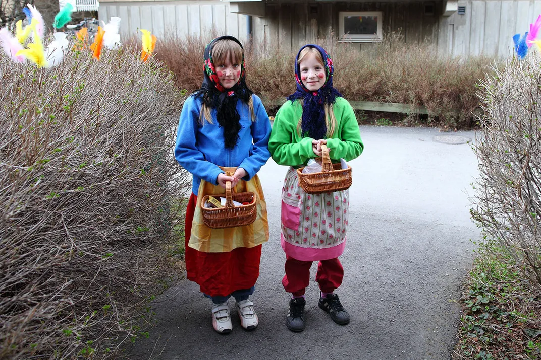 Two kids pose, dress in bright sweatshirts, black scarves, sneakers, and layers of skirts and aprons, each holding a basket. The bare bushes on either side of them are decorated with brightly colored artificial feathers.