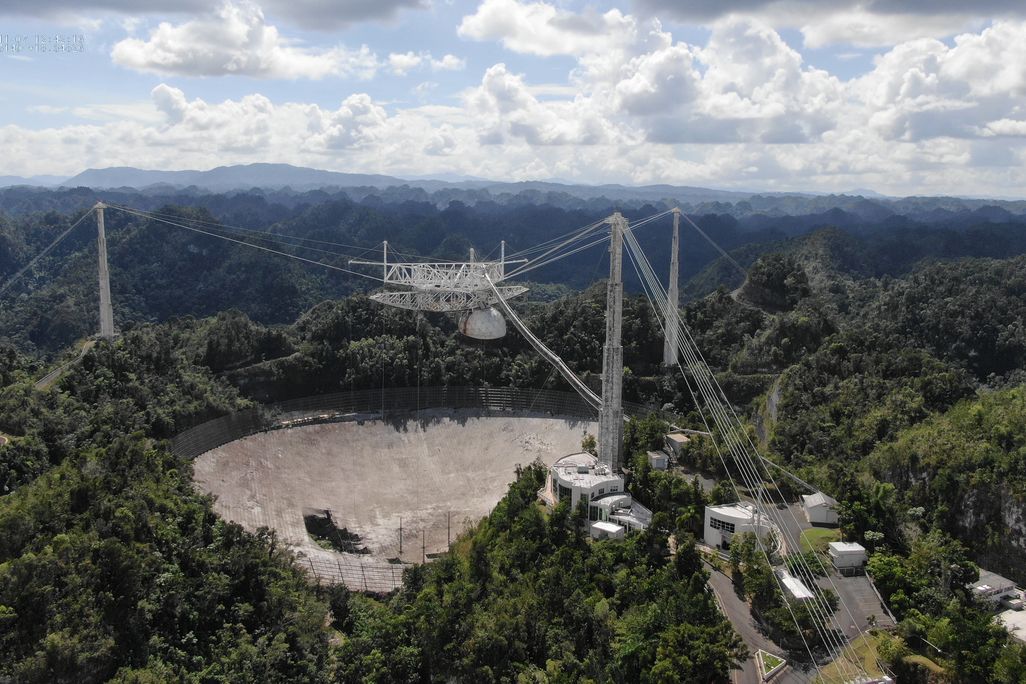A photograph of the dish from high above. It has three tall towers around the circular dish, all connected by thick cables. The dish has a 100-foot hole on the side. The observatory is surrounded by expansive forest. 