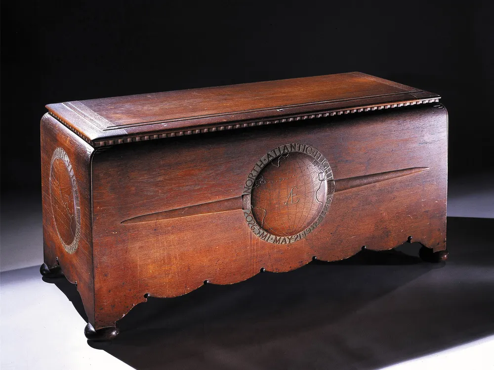A large hand-carved teakwood chest with scalloped edges and four round feet. The chest appears to be stained a rich reddish brown. The front panel on the chest has a bas-relief globe pattern flanked by a pair of airplane wings.