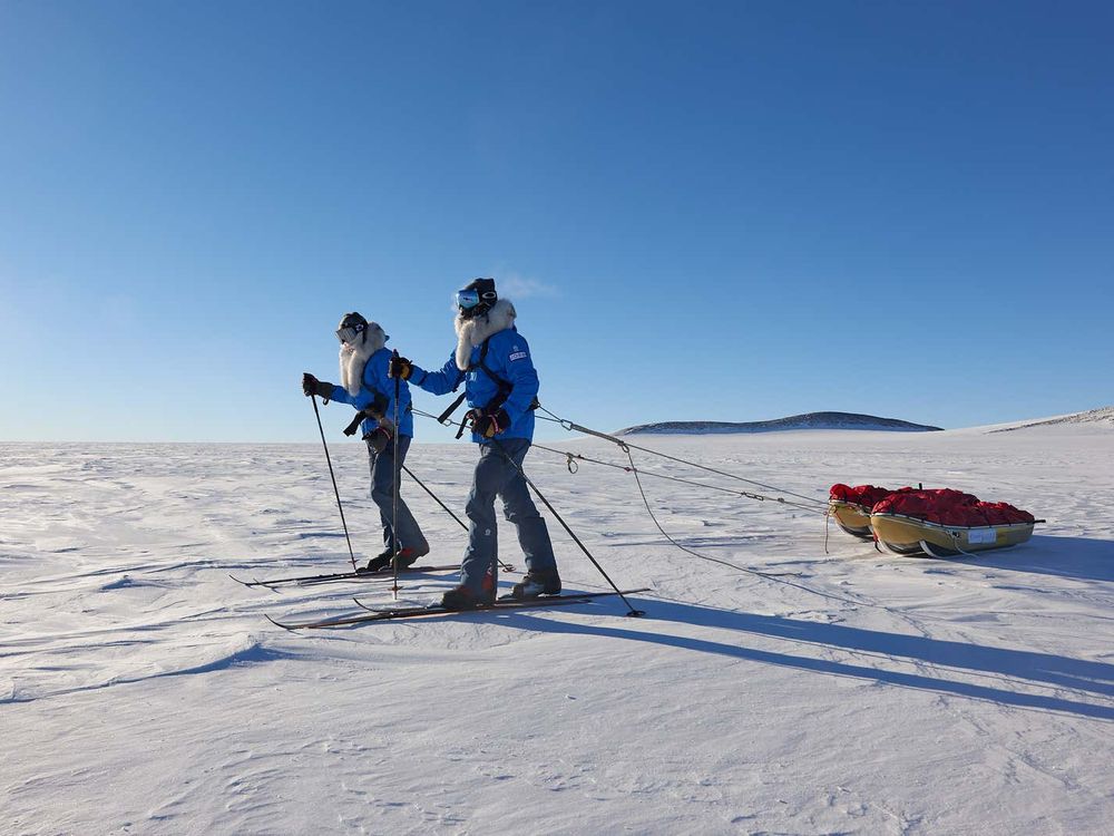 Two men in winter clothes and skis pull sleds across and frozen landscape