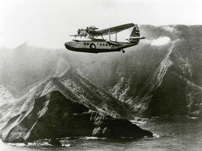 An Inter-Island Airways Sikorsky S-43 flies past Molokai mountains and lagoon.