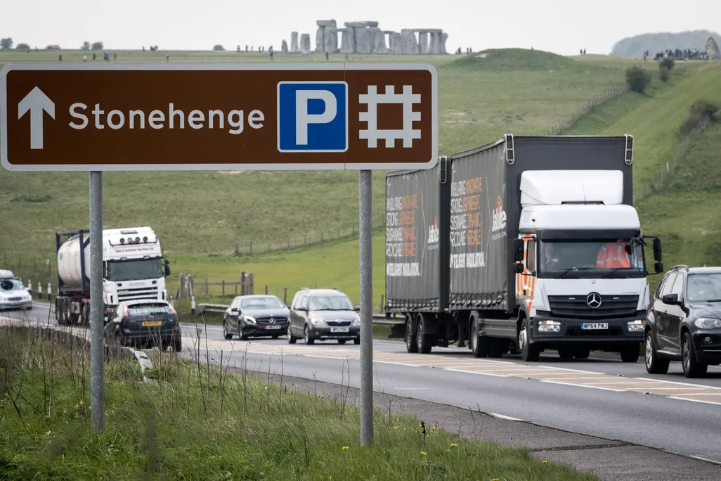 A busy road is in the foreground, with trucks and cars, and a large sign pointing to Stonehenge; the iconic stone structures are visible in the background
