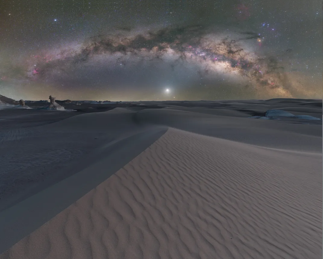 the milky way appears as an arc over the desert with dunes pointing up at the middle of the sky, where just above the horizon is a glowing white sphere