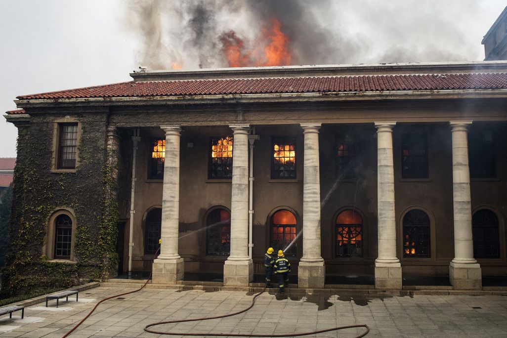 Two firefighters in gear, looking small compared to tall white columns, aim jets of water at the windows of a historic building; orange flames leap from the windows and ceiling of the structure and smoke pours out its top
