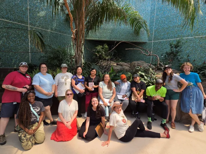 A group of teachers sits in two rows along a bench and on the ground with rocks and trees from the Bird House exhibit in the background.