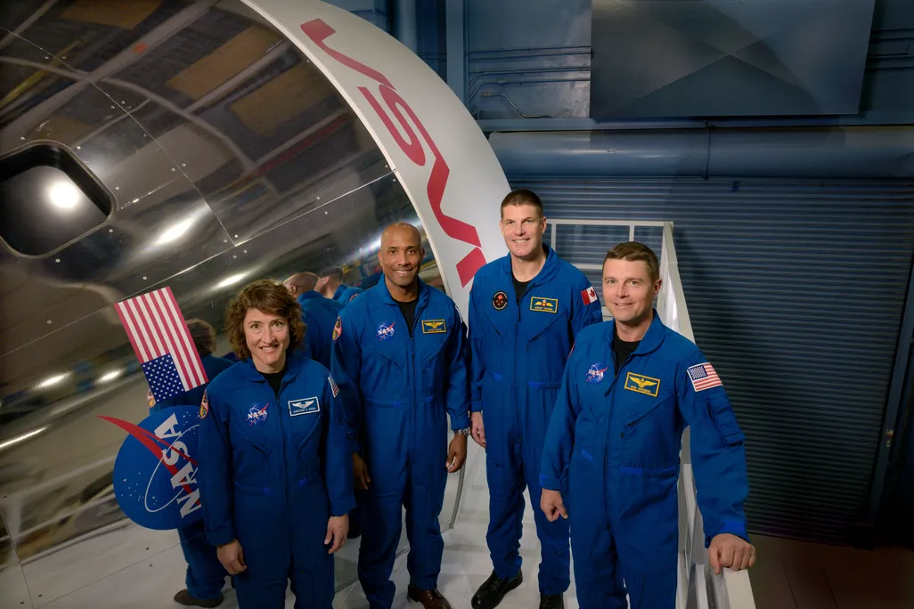 Four astronauts stand smiling in blue suits