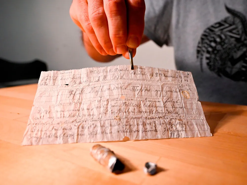 A person sits behind a table and delicately lifts the frail-looking slip of paper with tweezers; the sheet is thin and covered in spidery German cursive script; a small metal capsule that held the message lies on the table in front