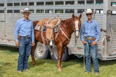 Two men in blue shirts, blue jeans, leather belts, and white cowboy hats pose in front of a brown horse.