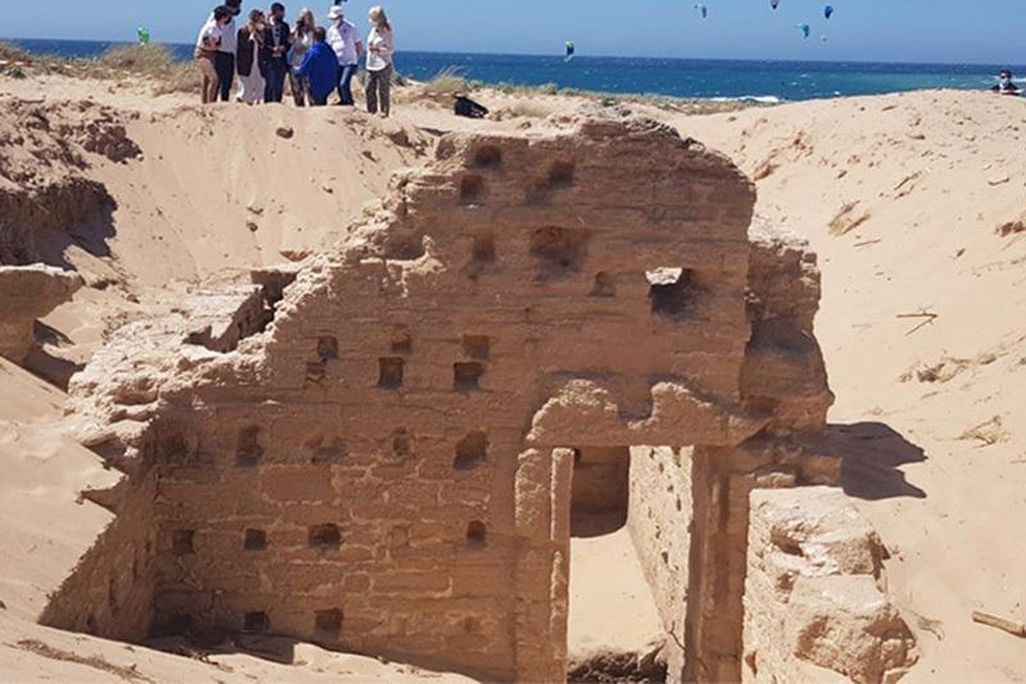 A group of people stand in front of a blue sky and blue sea while looking at a structure, with many small holes like windows and tall walls