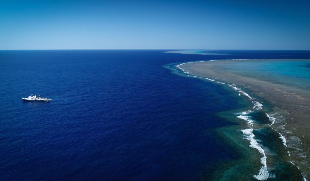 A photo taken from high above (likely by drone) shows the vast, bright blue ocean. The research vessel is on the left side. A long green and beige stretch of shallow coral reefs is on the right side. The horizon, where the deep blue ocean meets the lighte