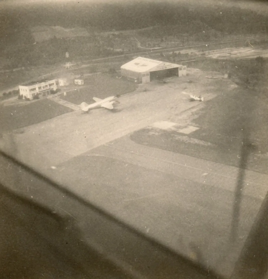 A black and white fuzzy aerial shot of an airplane hangar with a small white airplane parked on the runway
