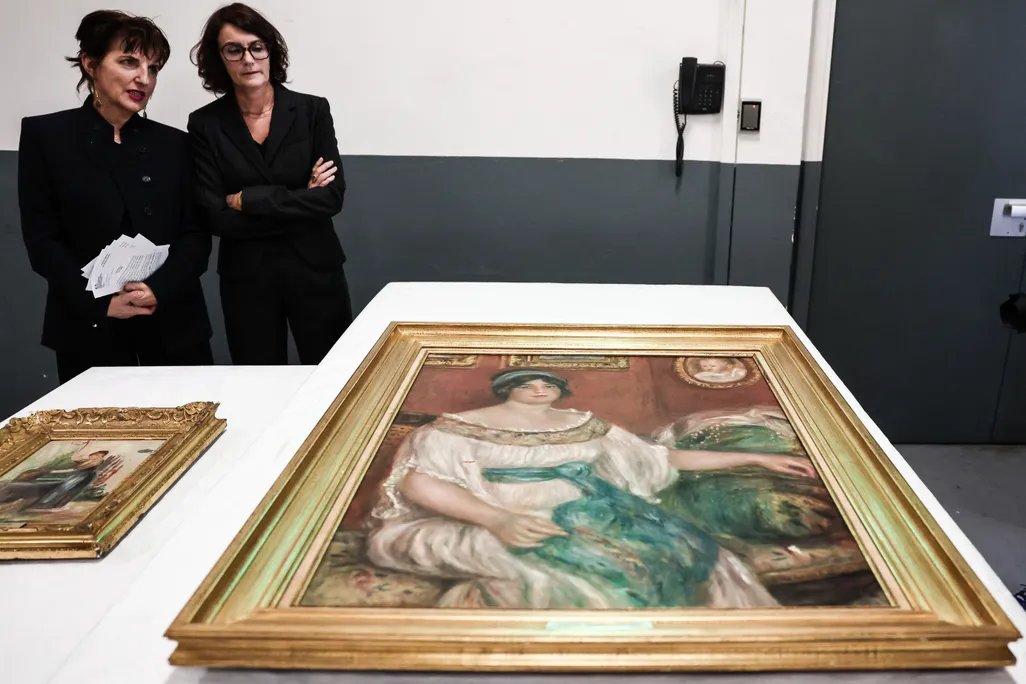 Two paintings on a table with two brunette women wearing black suits standing behind them against a wall