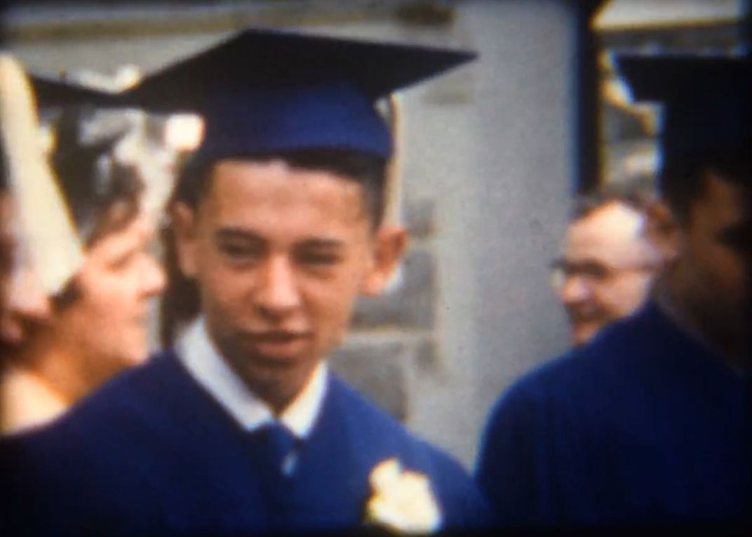 Old film still of young man in blue graduation cap and gown.