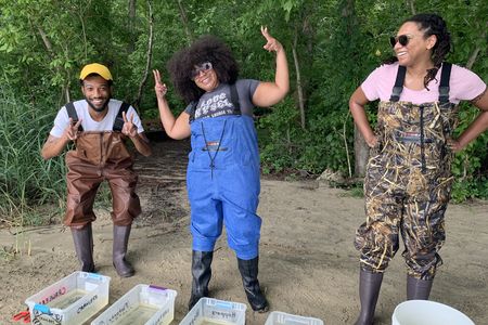 Three adults wearing chest waders laughing and in silly poses. They are standing on sand behind a line of plastic bins filled with water.
