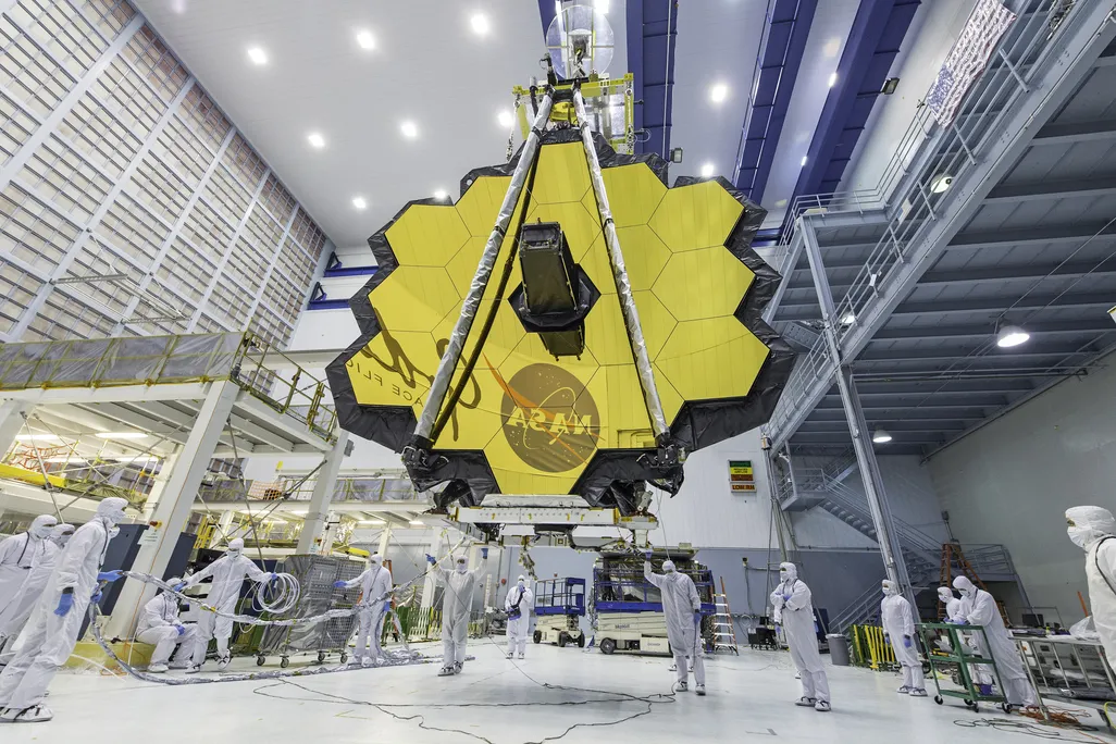 An image of the James Web Telescope in a NASA laboratory. Technicans are seen standing under it while the honey come shaped telecope mirror hangs above. The NASA logo is seen reflected in the mirrors.