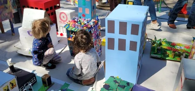Two tots kneel in the middle of several cardboard structures. They look into a colorful cardboard building filled with miniature sweets including donuts and cupcakes.