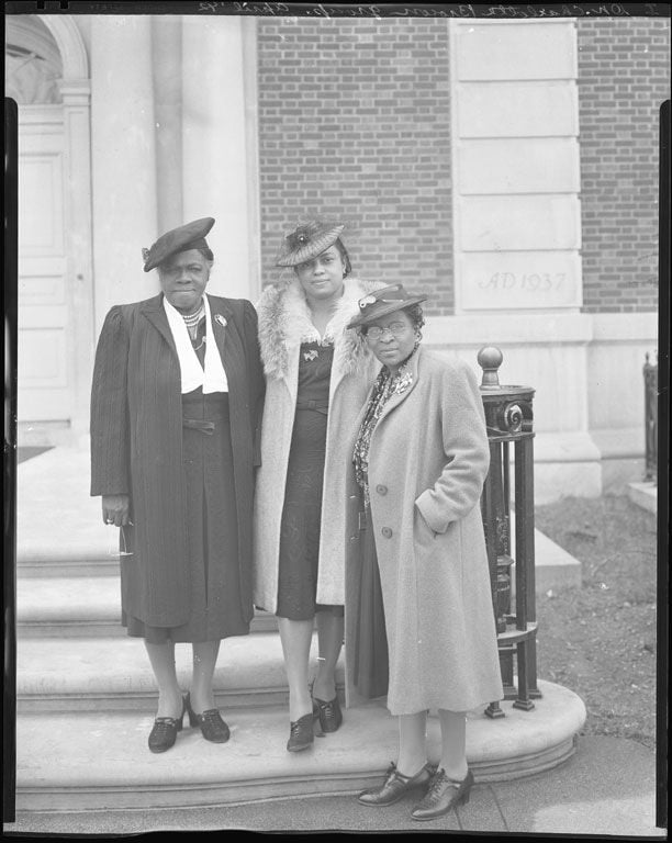 A black and white photograph of three African American women standing on the steps of a building. They all wear coats, hats, broaches, and stockings. The woman on the far left stares down the camera with determination.