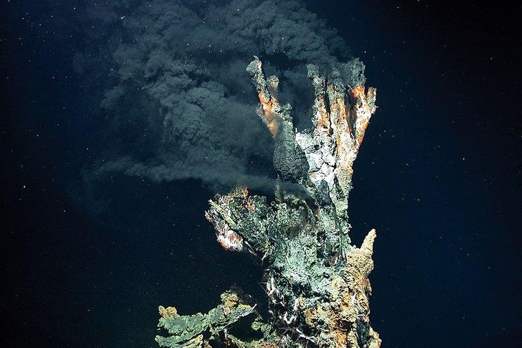 A hydrothermal vent in the deep ocean produces a dark, cloud of hot air against a dark blue background. The vent in the foreground is rocky, like a conglomerate of shells and rocks piled on top of each other.  