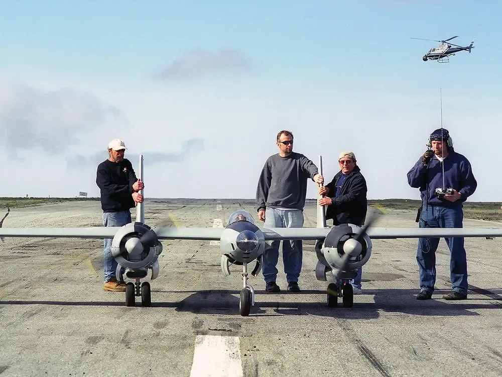Four men stand on a paved runway behind a large, grey twin-engine remote-controlled model airplane. The man on the far right holds a radio transmitter with a long antenna, while a small helicopter is visible in the sky behind him. The scene is set under