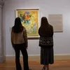 Two visitors stand in front of a colorful abstract painting and wall text in a museum gallery.